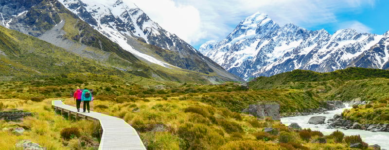 Couple walking along a scenic path through a valley with mountains in the background.